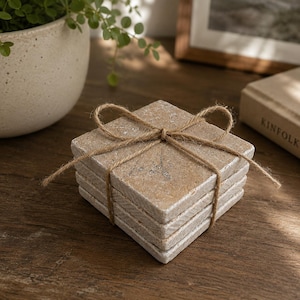 May include: A stack of four square stone coasters with a natural tan and grey mottled texture. The coasters are bound together with a thin brown jute twine tied in a bow on the top surface, creating a neat bundle of square drink protectors.