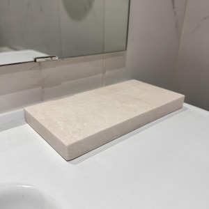 May include: A rectangular, light beige stone soap dish. It sits on a white countertop, with a mirror and white wall behind it. The soap dish has a simple, minimalist design, suitable for a bathroom.