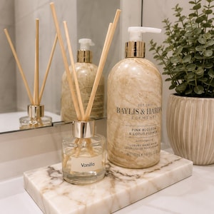 May include: A clear glass reed diffuser with a label reading Vanilla, a large beige marble-patterned bottle of Baylis & Harding Pink Blossom & Lotus Flower hand wash, and a rectangular marble tray, all arranged together on a white surface.