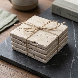 May include: A stack of four square, beige travertine stone coasters with a porous, natural texture. The coasters are tied together with a thin, brown jute twine string tied in a bow on the top surface of the uppermost coaster in the stack.