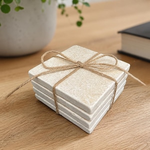 May include: A stack of four square, off-white stone coasters with a textured, mottled surface. The coasters are bound together by a thin, brown jute twine tied in a bow across the top centre, securing the set in a neat, uniform arrangement.