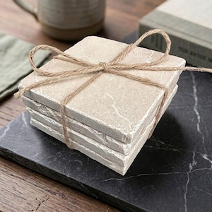 May include: A stack of four square beige stone coasters with natural veining, tied together with a thin brown jute twine bow. The coasters have a rough, textured edge and a smooth top surface, presented as a bundled set for table protection.