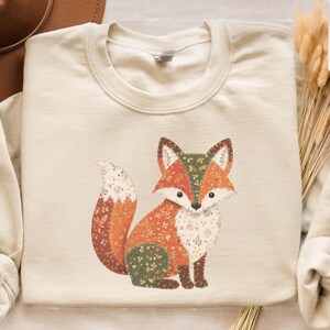 May include: A sand-colored sweatshirt featuring a colorful fox illustration. The fox design incorporates shades of orange, green, and white. The sweatshirt has long sleeves and a crew neck. The brand name "Gildan" is visible in the bottom left corner.