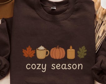 Könnte beinhalten: Dunkel schokoladenbraunes Sweatshirt mit Rundhalsausschnitt. Auf der Vorderseite befindet sich ein gesticktes Design aus Ahornblatt, Kaffeetasse, Kürbis, Kerze und Blatt mit den Worten "cozy season" darunter. Die Marke Gildan ist sichtbar.