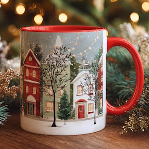 May include: A ceramic mug with a red interior and handle. The exterior features a winter village scene with red, green, and white houses, bare trees, and string lights. The mug is labeled "11oz".