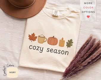 Könnte beinhalten: Cremefarbenes T-Shirt mit einem herbstlichen Motiv. Das Design umfasst ein Ahornblatt, eine Tasse Schlagsahne, einen Kürbis, eine Kerze und ein Eichenblatt. Darunter steht der Schriftzug "cozy season".