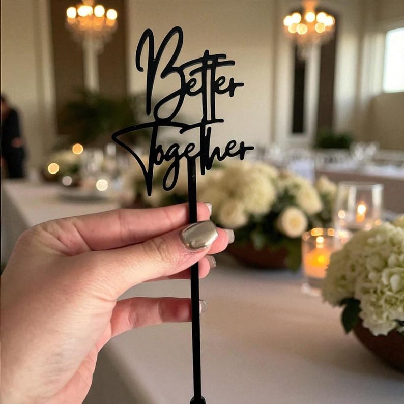 Pu&ograve; includere: Un cake topper nero con le parole "Better Together" in corsivo. Il topper &egrave; tenuto davanti a una tavola imbandita con fiori bianchi e candele, suggerendo un tema di matrimonio o evento.