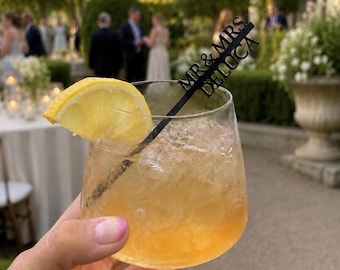 May include: A clear glass filled with an amber-colored liquid, ice cubes, and a yellow lemon slice garnish. A black acrylic drink stirrer is inserted into the glass, featuring the laser-cut text MR & MRS DELUCA in a bold, sans-serif font.