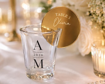 May include: A clear glass shot glass featuring a black printed monogram with the letters A and M separated by the year 2026. A circular gold-toned acrylic place card is slotted into the rim, displaying the text TABLE 1 and the name Olivia.