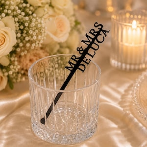 May include: A clear, vertical-ribbed glass tumbler containing a black acrylic drink stirrer. The stirrer features the laser-cut text MR & MRS DELUCA oriented diagonally along its length, with a circular base at the bottom of the glass.