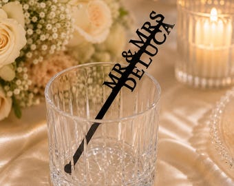 May include: A clear, vertical-ribbed glass tumbler containing a black, custom-cut acrylic drink stirrer. The stirrer features the text "MR & MRS DELUCA" in a bold, sans-serif font, extending diagonally from the base of the glass to the top rim.