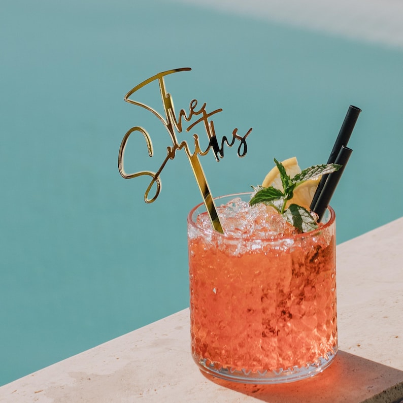 Pu&ograve; includere: Un cocktail in un bicchiere strutturato con ghiaccio, menta e una fetta di limone. Un topper dorato con le parole "The Smiths" &egrave; nella bevanda. Due cannucce nere sono nella bevanda. Lo sfondo &egrave; azzurro.