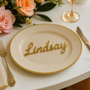 May include: A cream-colored plate with a gold rim features the name "Lindsay" in elegant gold script. A gold fork and knife are placed on the table, alongside a wine glass and floral arrangement. This is a place setting for a special event.