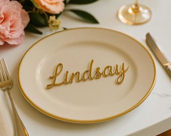 May include: A cream-colored plate with a gold rim features the name "Lindsay" in elegant gold script. A gold fork and knife are placed on the table, alongside a wine glass and floral arrangement. This is a place setting for a special event.