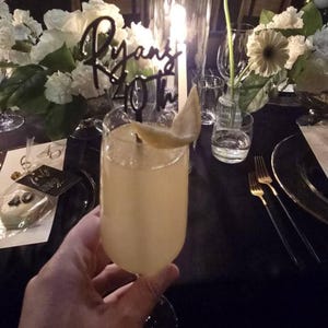 Pu&ograve; includere: Un cocktail in un bicchiere alto con una fetta di limone, davanti a una tavola. Un cartello nero con la scritta "Ryan's 40th" &egrave; sullo sfondo. La tavola &egrave; apparecchiata con fiori, candele e forchette dorate.
