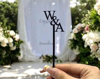 May include: A black acrylic drink stirrer featuring the letters W and A separated by an ampersand symbol. The topper is designed with a serif font and is attached to a long, thin, straight stem that ends in a small circular base at the bottom.