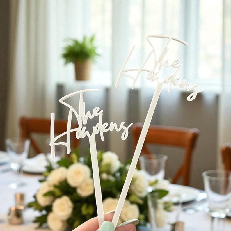 Pu&ograve; includere: Due decorazioni da tavolo bianche con la scritta "The Haydens" in corsivo. Le decorazioni sono tenute davanti a una tavola con fiori bianchi e vetreria. Una pianta in vaso &egrave; visibile sullo sfondo.