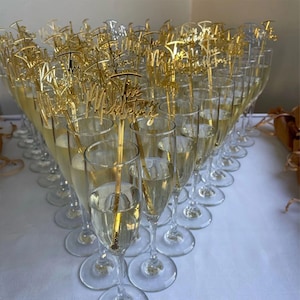 Pu&ograve; includere: Fl&ucirc;te di champagne piene di una bevanda frizzante, ognuna ornata da un topper dorato con la scritta "The Wedding". I bicchieri sono disposti su una superficie bianca, creando un'atmosfera festosa per un matrimonio.