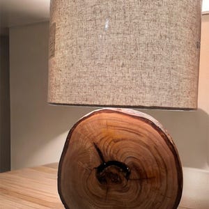 Puede incluir: Lámpara de mesa de madera con base de madera natural y pantalla de tela beige. La base es una sección transversal de un tronco de árbol, mostrando la veta de la madera y un centro oscuro. La pantalla es cilíndrica.