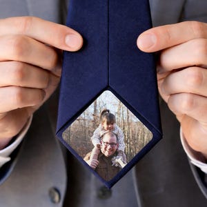 May include: Navy blue tie with a photo insert. The photo shows a man with glasses and a young girl on his shoulders, set against a backdrop of trees. The tie is held up by a person wearing a gray suit.