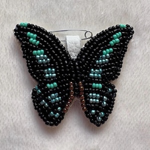 May include: A black and turquoise beaded butterfly brooch with a silver pin and white leather backing.