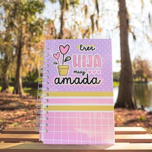 Op de afbeelding: Een spiraalgebonden notitieboek met een lavendelkleurige kaft met de tekst "Eres Hija Muy Amada" en een bloemmotief. Het notitieboek heeft roze, gele en witte gestreepte accenten en een roze rasterpatroon aan de onderkant.