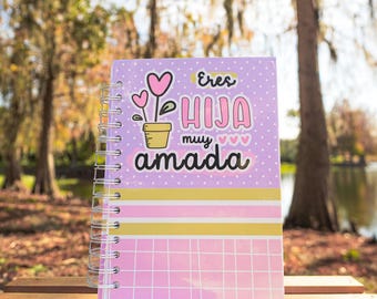 May include: A spiral-bound notebook with a lavender cover featuring the text "Eres Hija Muy Amada" and a floral design. The notebook has pink, yellow, and white striped accents and a pink grid pattern on the bottom.