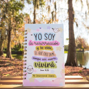 May include: A pink and blue devotional journal with a white background and a quote in Spanish that reads "YO SOY la resurrección y la vida. EL QUE CREE EN MI aunque esté muerto, vivirá." The quote is from John 11:25. The journal has a gold spiral binding and a white sheep illustration.