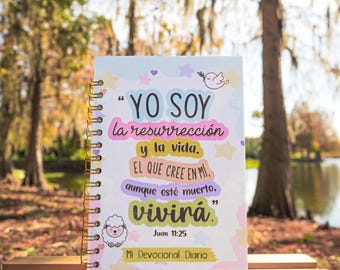 May include: A pink and blue devotional journal with a white background and a quote in Spanish that reads "YO SOY la resurrección y la vida. EL QUE CREE EN MI aunque esté muerto, vivirá." The quote is from John 11:25. The journal has a gold spiral binding and a white sheep illustration.