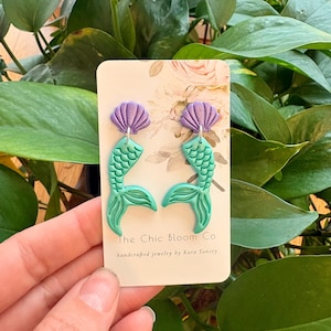 May include: A pair of handcrafted mermaid earrings. The earrings feature a purple seashell top and a turquoise mermaid tail design. The card behind the earrings reads "The Chic Bloom Co" and "handcrafted jewelry by Kara Yancey".