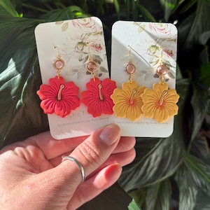 May include: Two pairs of handcrafted hibiscus flower earrings. One pair is red, the other is yellow. Each earring has a gold-colored hook and a small, round, pink bead. The earrings are displayed on floral-patterned cards.