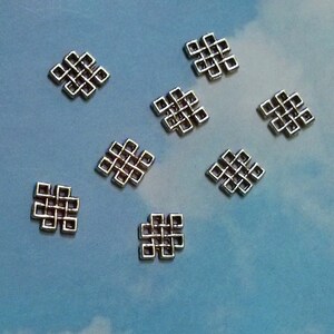May include: Eight silver-toned metal charms in the shape of a square knot, also known as an endless knot. The charms are arranged in a scattered pattern on a light blue background.