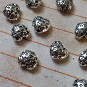 May include: A group of silver heart-shaped beads with a textured surface. The beads have a hole in the top for stringing.