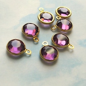 May include: Close-up of several purple gemstone pendants with gold-colored metal settings. The gemstones are round and faceted, with a gradient of color from light to dark purple. Each pendant has a small loop for attaching to a chain or other jewelry component.