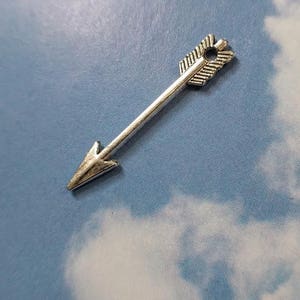 May include: Silver arrow charm with a detailed arrowhead and fletching. The arrow is pointing to the right.
