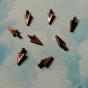 May include: Eight antique copper-toned arrowhead-shaped charms with a loop at the top. The charms are about 1 inch long and have a textured surface.