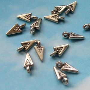 May include: Small, silver-colored arrow charms with a textured arrowhead and a loop for attaching to jewelry. The charms have a vintage or antique appearance, with a slightly tarnished finish. The arrows are scattered on a light blue surface.