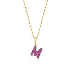 May include: A gold-coloured necklace featuring a pendant shaped like the letter "M". The letter is set with small, bright pink gemstones. The necklace is displayed against a white backdrop.