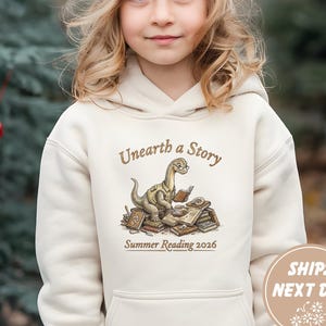 Puede incluir: Una sudadera con capucha color crema con un dinosaurio leyendo libros, con el texto "Unearth a Story" y "Summer Reading 2026". La sudadera con capucha la lleva una persona joven con un gorro beige.