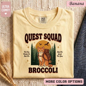 May include: A light yellow t-shirt with the text "QUEST SQUAD BROCCOLI" in brown. The shirt features a cartoon cat with a backpack in front of a forest and a sun. The shirt also has the text "No One Wants It, But It's" and "But It's Good For You".