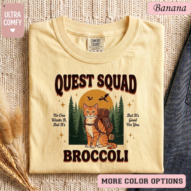 May include: A light yellow t-shirt with the text "QUEST SQUAD BROCCOLI" in brown. The shirt features a cartoon cat with a backpack in front of a forest and a sun. The shirt also has the text "No One Wants It, But It's" and "But It's Good For You".