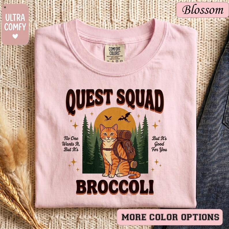May include: A light pink t-shirt with the words "QUEST SQUAD BROCCOLI" in brown lettering. The shirt features a cartoon cat with a backpack in front of a forest scene. The text "No One Wants It, But It's Good For You" is also printed on the shirt.