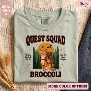 May include: A light green t-shirt with the words "QUEST SQUAD BROCCOLI" in brown lettering. The shirt features a cartoon cat with a backpack in front of a forest scene. The text "No One Wants It, But It's Good For You" is also included.