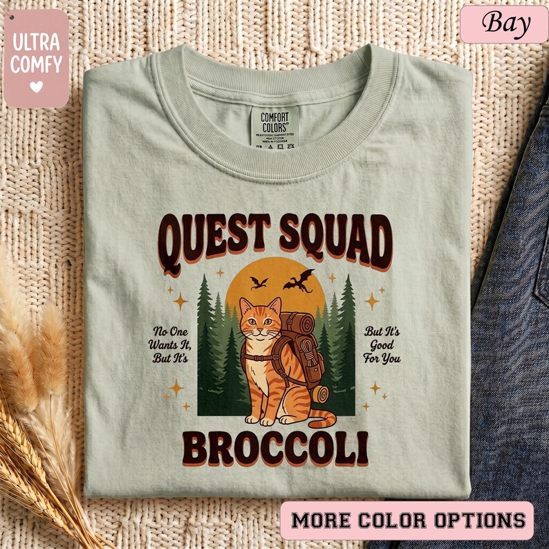 May include: A light green t-shirt with the words "QUEST SQUAD BROCCOLI" in brown lettering. The shirt features a cartoon cat with a backpack in front of a forest scene. The text "No One Wants It, But It's Good For You" is also included.