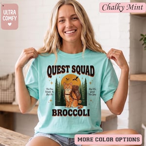 May include: A light blue t-shirt with the words "QUEST SQUAD" in a brown, distressed font. The shirt features a cartoon cat with a backpack in front of a forest scene. Text on the shirt includes "No One Wants It, But It's Good For You" and "BROCCOLI".