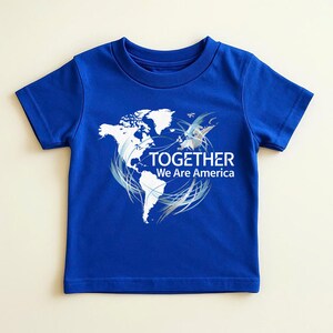 Puede incluir: Una camiseta azul real con un contorno blanco de las Am&eacute;ricas y la frase "TOGETHER We Are America". El dise&ntilde;o incluye remolinos plateados y un gr&aacute;fico estilizado de un p&aacute;jaro. La camiseta est&aacute; hecha de un material suave.