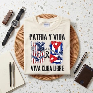 Puede incluir: Camiseta color crema con las frases "PATRIA Y VIDA" y "VIVA CUBA LIBRE" en negro. El diseño incluye las banderas estadounidense y cubana, águilas, la Estatua de la Libertad, una cadena y una cinta negra.