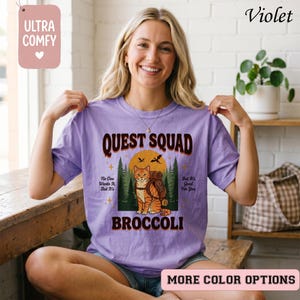 May include: A violet-colored t-shirt with the text "QUEST SQUAD" and a graphic of a cat with a backpack in front of a sunset. The shirt also has the text "BROCCOLI" and "ULTRA COMFY" on it.