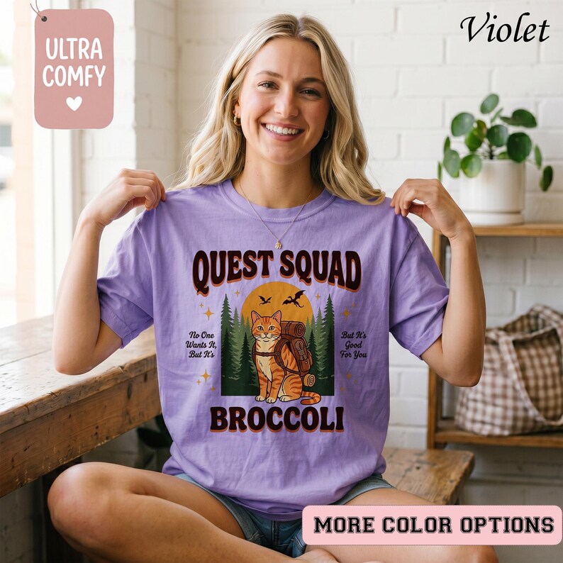 May include: A violet-colored t-shirt with the text "QUEST SQUAD" and a graphic of a cat with a backpack in front of a sunset. The shirt also has the text "BROCCOLI" and "ULTRA COMFY" on it.