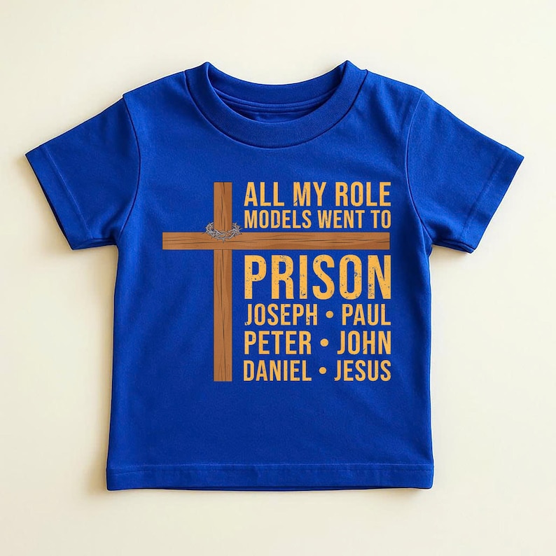 May include: A royal blue t-shirt with the text "ALL MY ROLE MODELS WENT TO PRISON" in yellow. Below the text are the names Joseph, Paul, Peter, John, Daniel, and Jesus. A brown cross graphic is on the left.